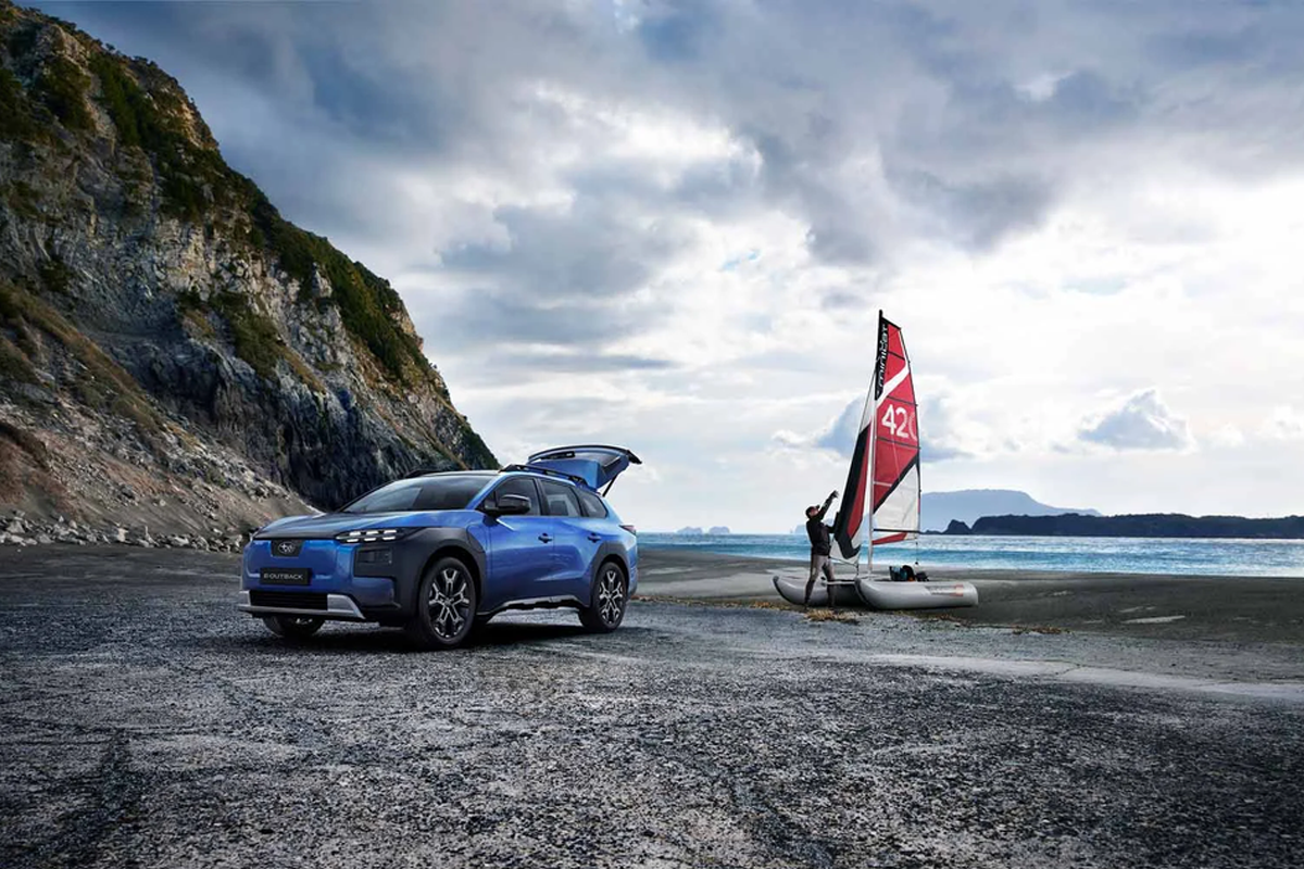 blauer E-Outback am Strand, daneben steht ein Mann mit einem Windsurfer